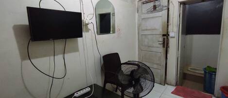 A small room with a television mounted on the wall, a power strip, a fan, a chair, and a door leading to another room.
