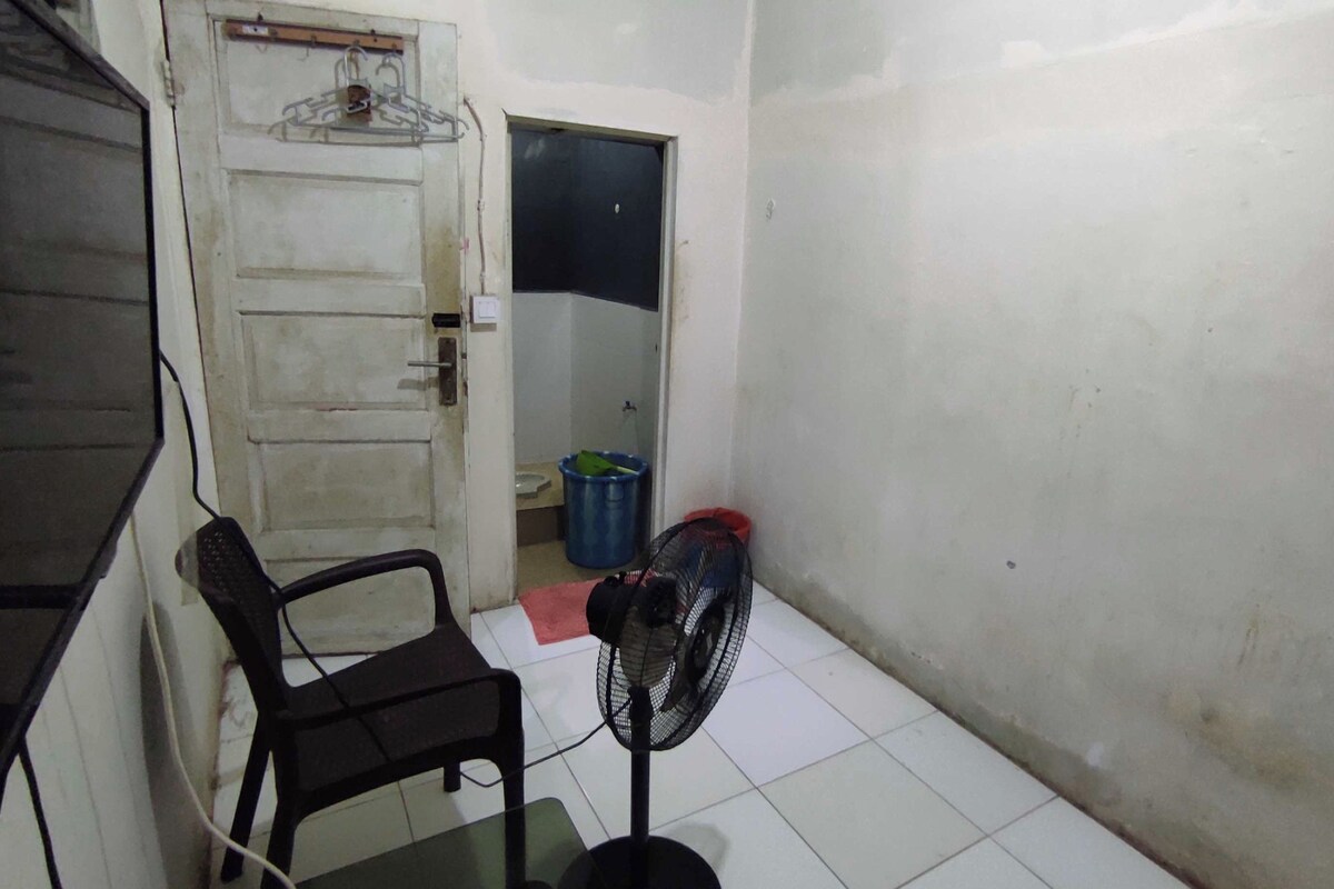 A small, dimly lit room with a white door, a black plastic chair, a standing fan, and a blue trash bin.