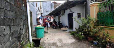 A narrow alleyway with a motorcycle parked on the right, a green trash bin on the left, and potted plants along the right side.