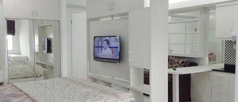 A bedroom with a bed, a TV mounted on the wall, a wardrobe, and a small kitchenette area.