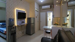 A compact living space with a kitchenette, a flat-screen TV mounted on the wall, a refrigerator, a water dispenser, a sofa, and a small table with a mirror. - Apartment Gading Nias Residence (Jakarta)