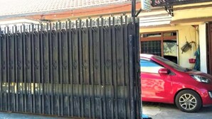 A red car parked in front of a black metal gate, with a tiled roof and a building with a yellow wall in the background. - Penginapan Meli (Surabaya)