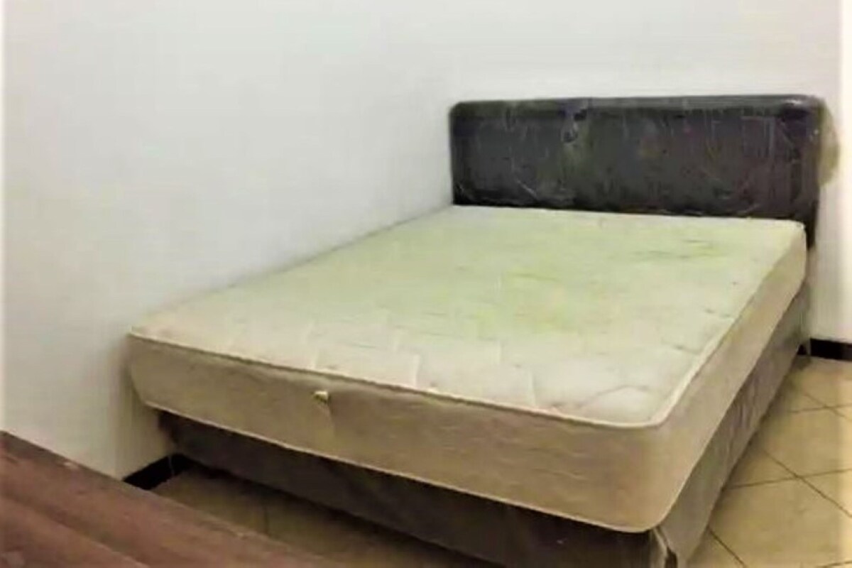 Pequeña habitación individual con una cama y colchón adosados a la pared, una puerta visible y suelo de baldosas.