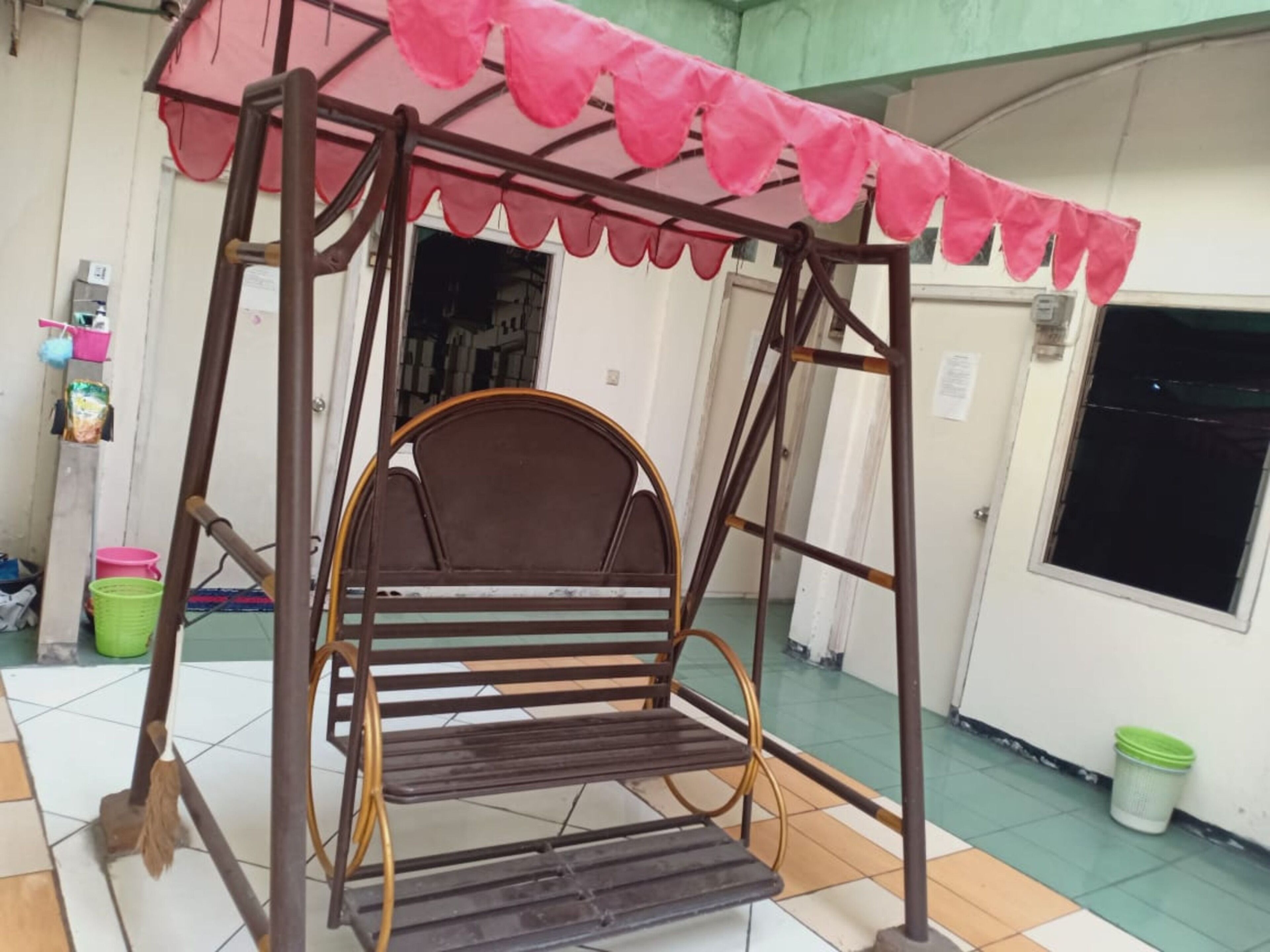 A swing chair with a metal frame and a pink canopy, situated in a room with tiled flooring and a door in the background.