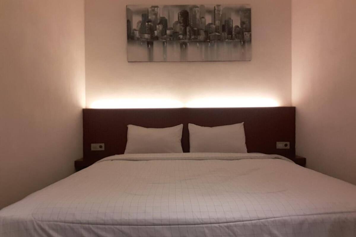 Une chambre d’hôtel avec un grand lit, une tête de lit dotée d’un éclairage intégré et un tableau représentant un paysage urbain accroché au mur.