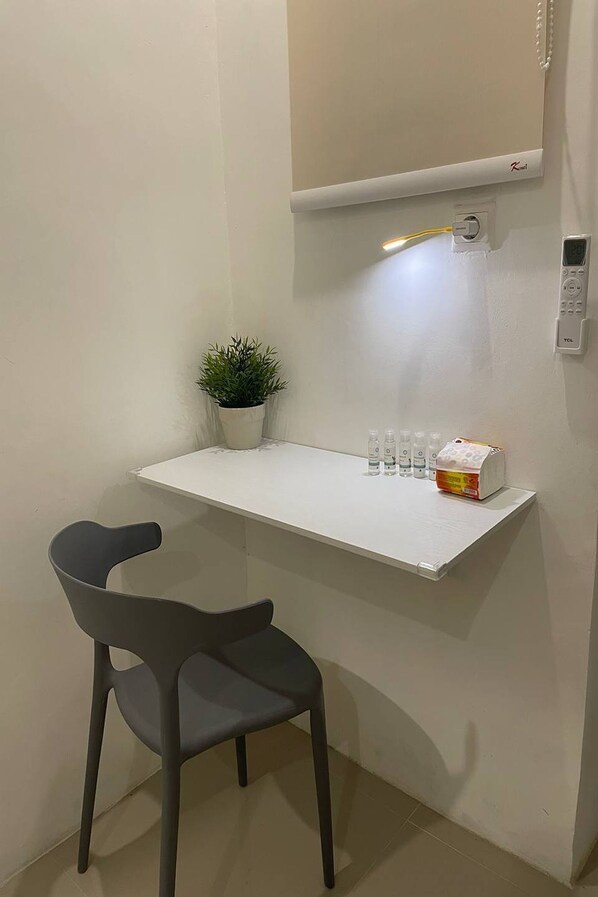 Una habitación pequeña y luminosa con un escritorio blanco, una planta en maceta, una silla y una estantería con botellas y una caja.