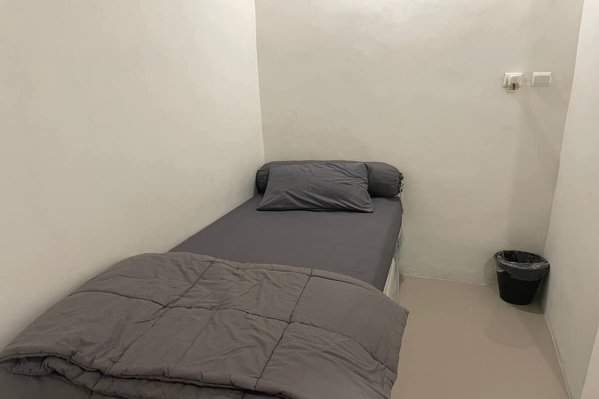 A small, single-bed room with a grey bedspread and pillow, a trash bin, and a light switch on the wall.