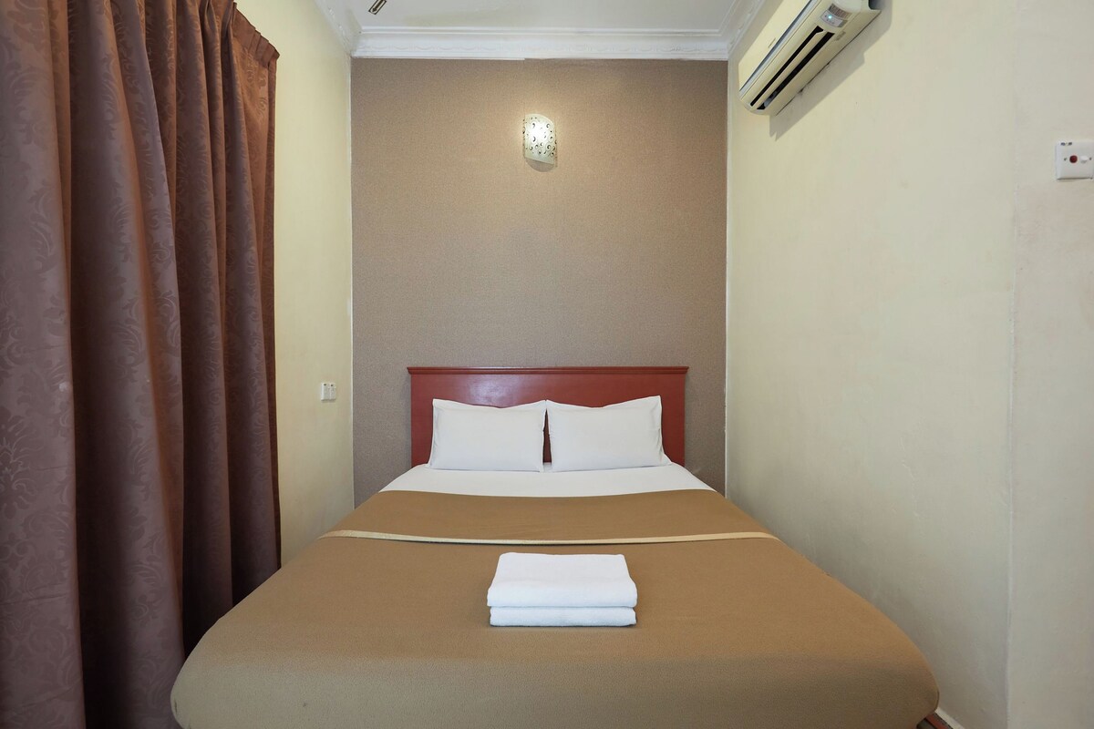 A neatly made bed with a brown bedspread and white pillows, a folded white towel on the bed, and a brown curtain partially drawn to the side.