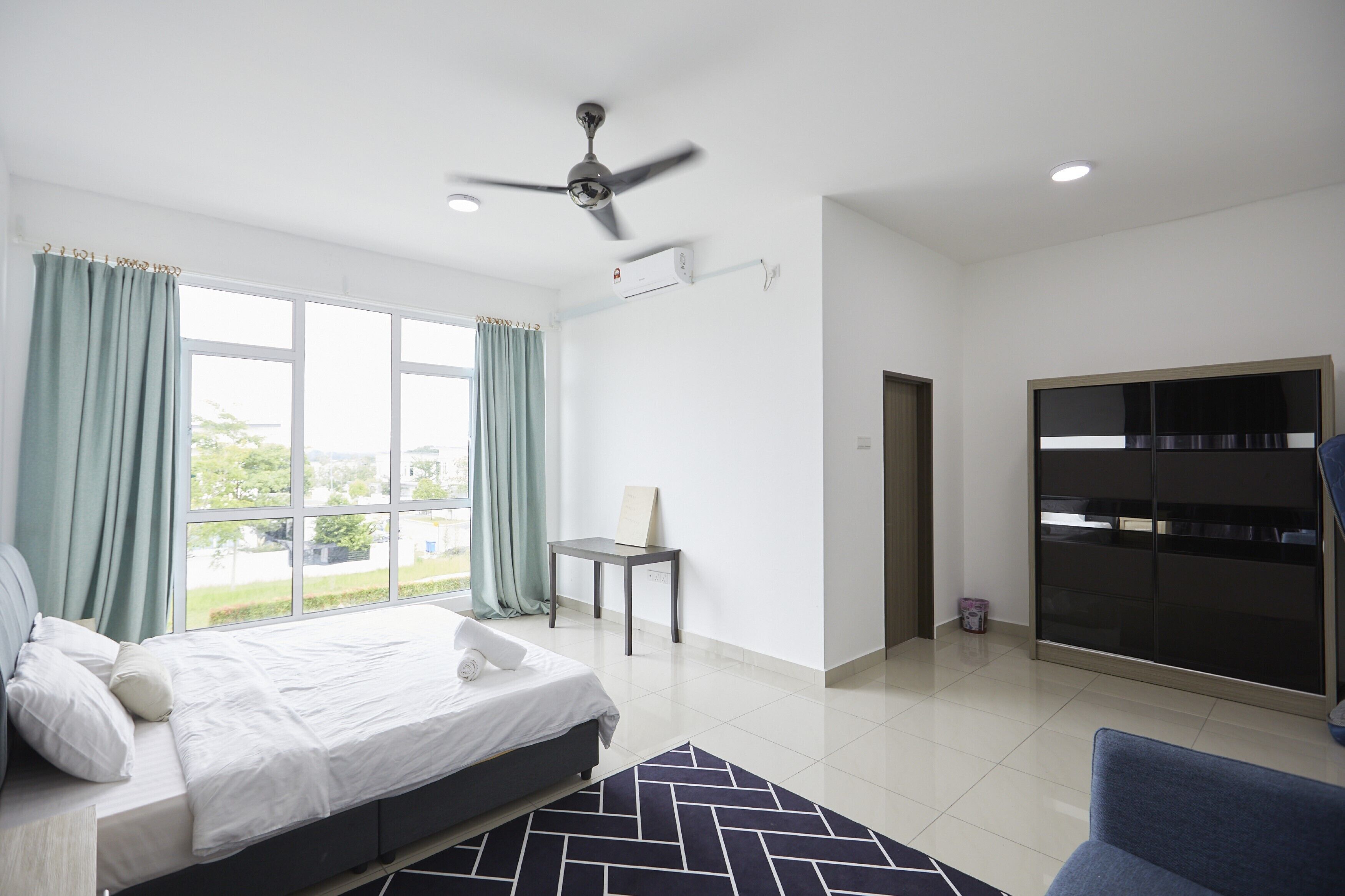 Habitación de hotel moderna con cama, escritorio, silla, ventilador de techo y un gran ventanal con cortinas.