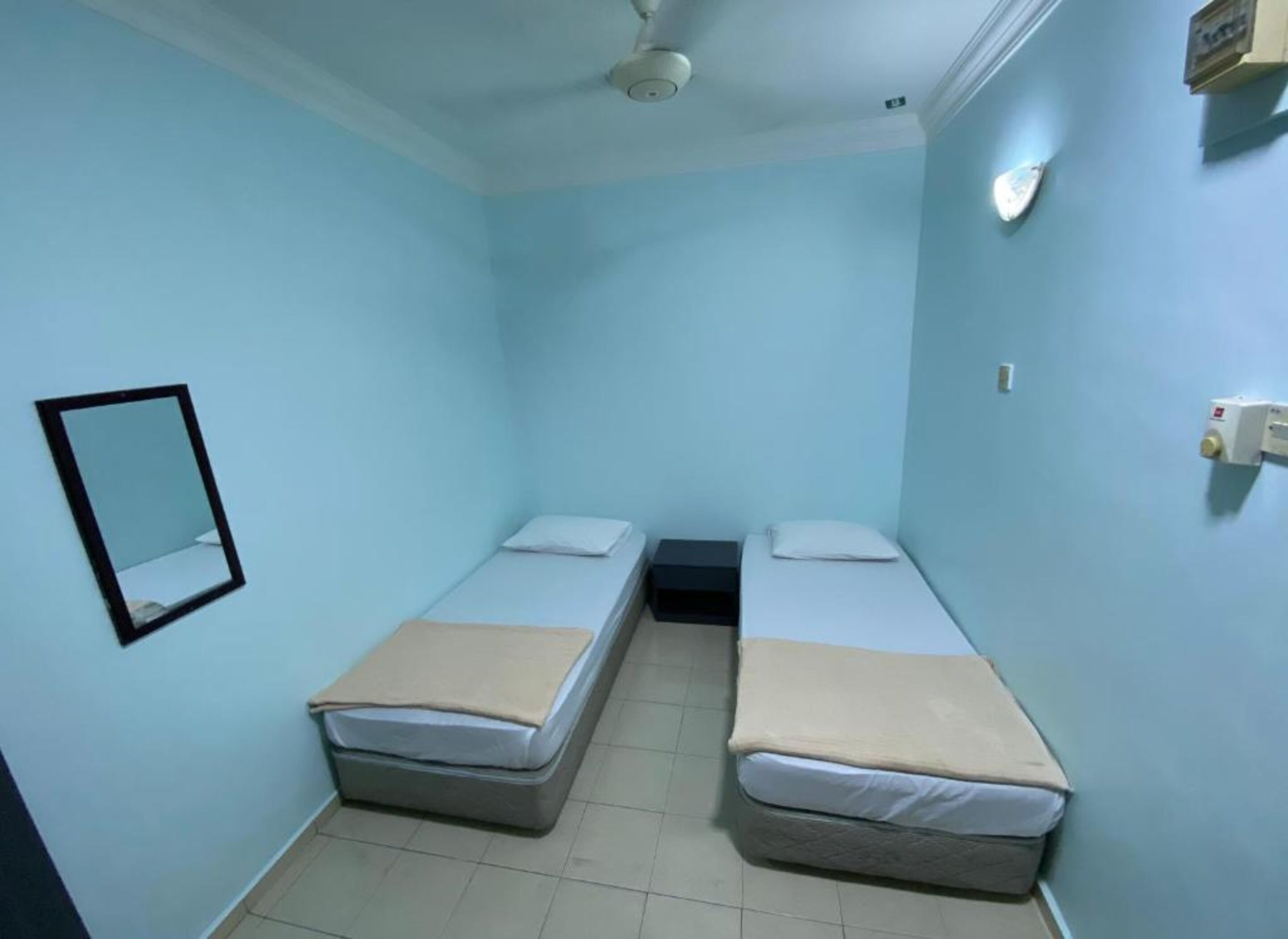 A small, single-room with two single beds, a ceiling fan, and a wall-mounted mirror.