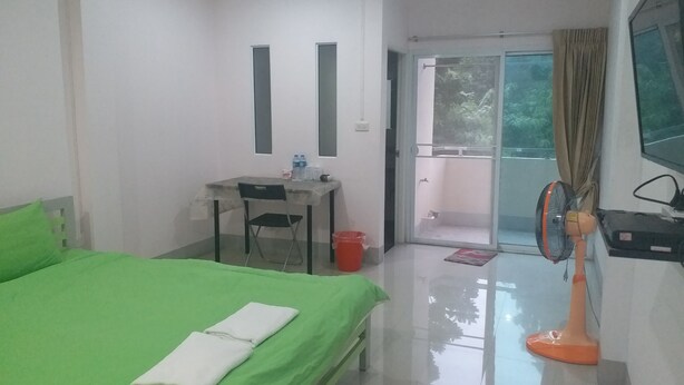 Habitación de hotel con una cama verde, un escritorio con silla, una mesita con botellas de agua, un ventilador y un televisor en la pared.