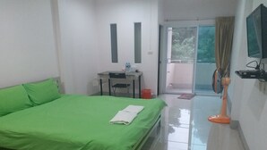 Habitación de hotel con cama verde, escritorio con silla, pequeña mesa con una botella, ventilador y televisión en la pared.