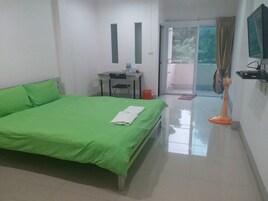 Habitación de hotel con cama verde, escritorio con silla, pequeña mesa con una botella, ventilador y televisión en la pared.