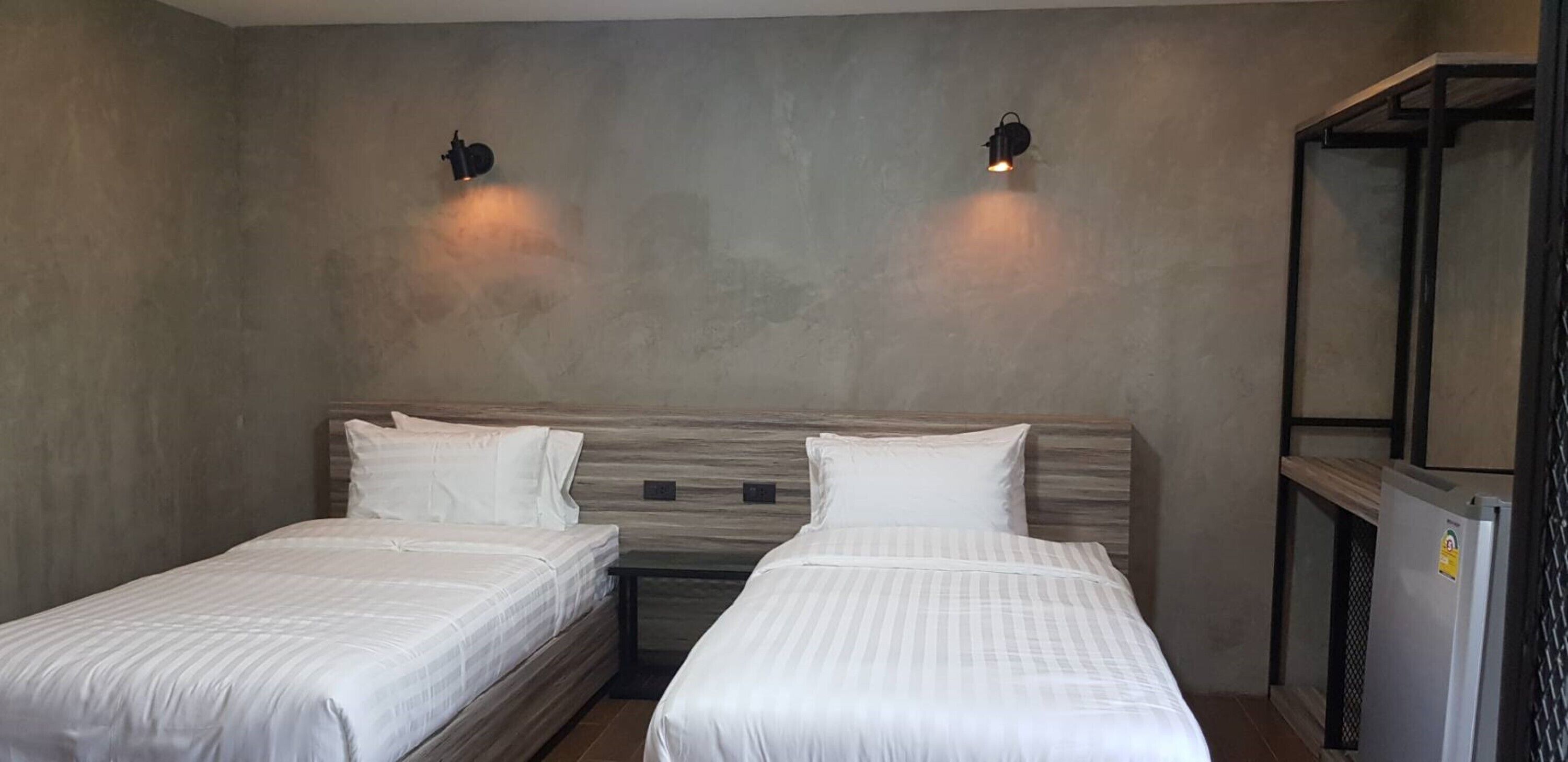 Habitación de hotel con dos camas individuales, cabecero de madera, una mesita y un refrigerador.