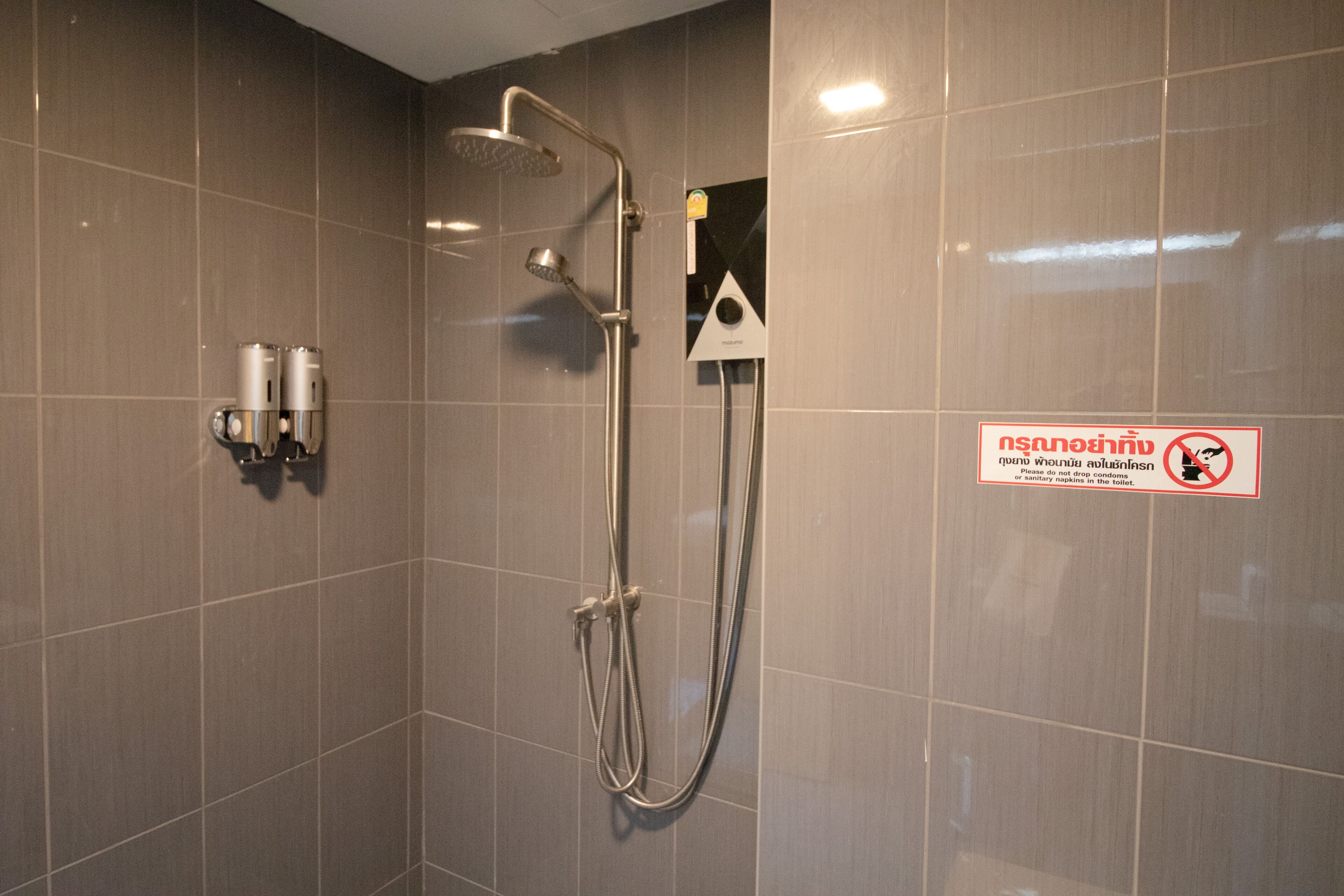 Zona de ducha con ducha de mano, cabezal de ducha portátil y grifo empotrado en la pared. Hay un cartel con un círculo rojo y una línea que indica prohibido secarse con toallas.