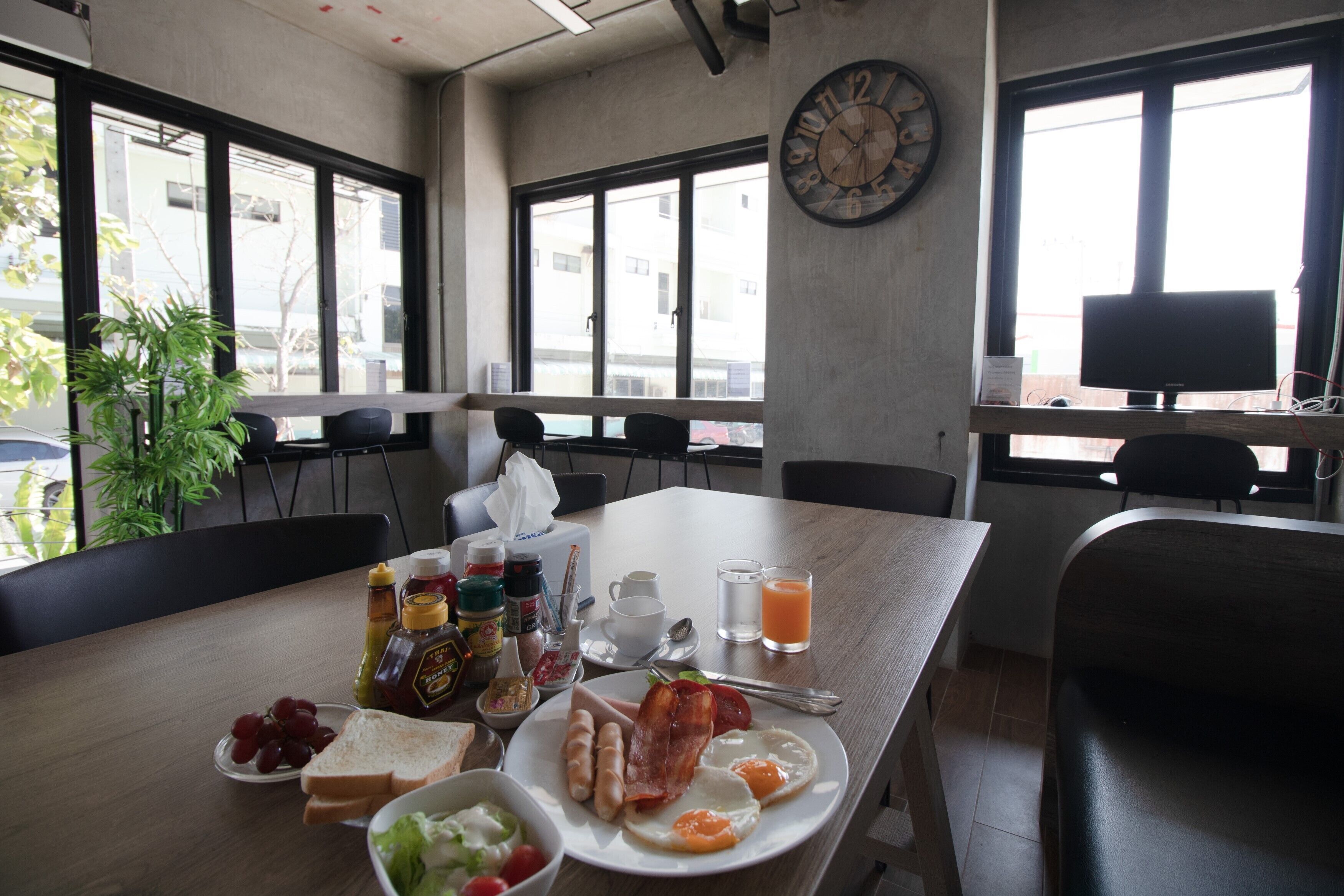 A breakfast table with eggs, bacon, toast, and fruit. There are chairs around the table, a clock on the wall, and a window with a view of buildings outside.