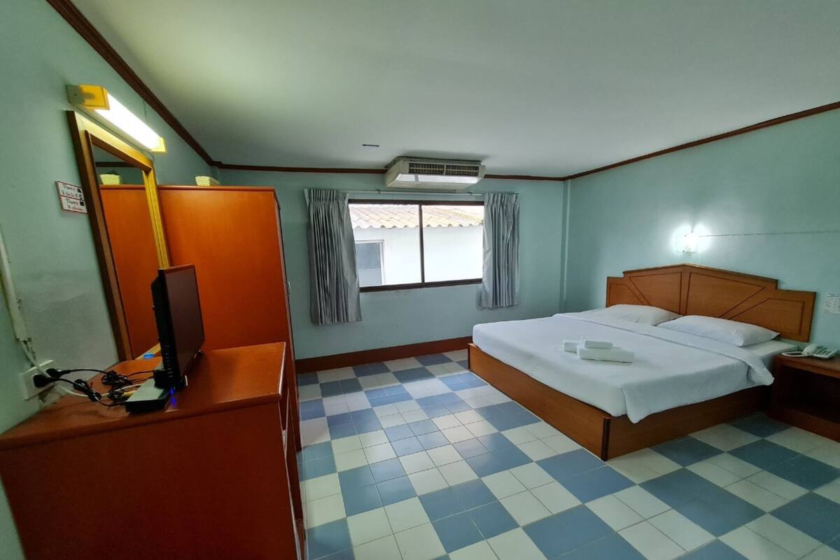 Une chambre d’hôtel avec un sol à carreaux, un lit avec du linge de maison blanc, une tête de lit en bois, une table de chevet avec un téléphone, un bureau avec un ordinateur et une fenêtre avec des rideaux.