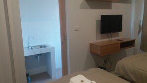 A bedroom with a bed, a TV mounted on the wall, a wooden shelf, and a bathroom visible through an open door. - Saney Apartment (Pak Chong)