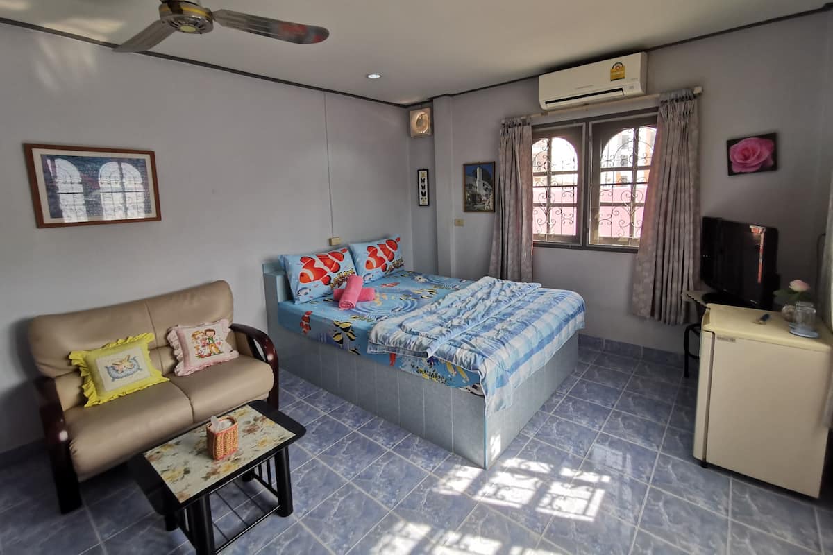 A bedroom with a bed, a sofa, a small table, a ceiling fan, an air conditioning unit, a TV, and a window with curtains.