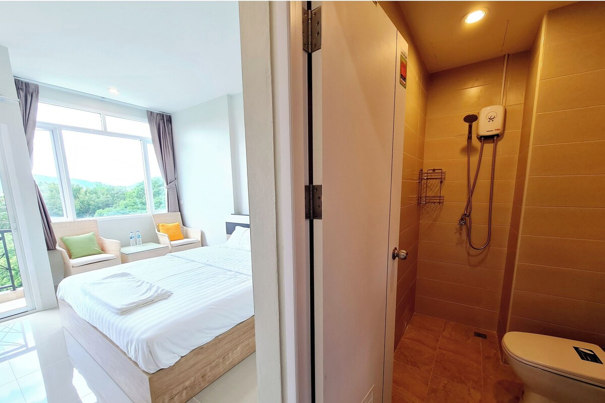 A hotel room with a large bed, a bathroom with a shower, and a view of greenery outside.