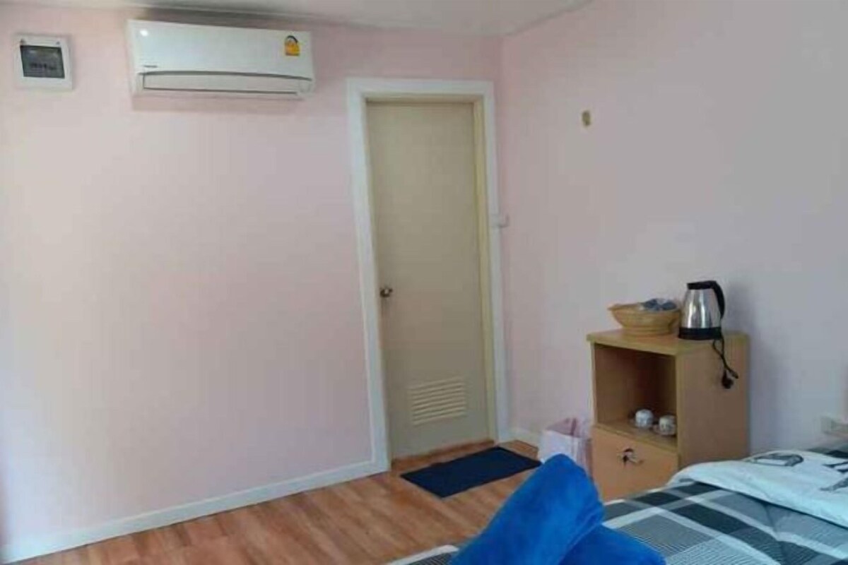 Una habitación con una cama, suelo de madera, una puerta, un estante pequeño con una tetera y un equipo de aire acondicionado.