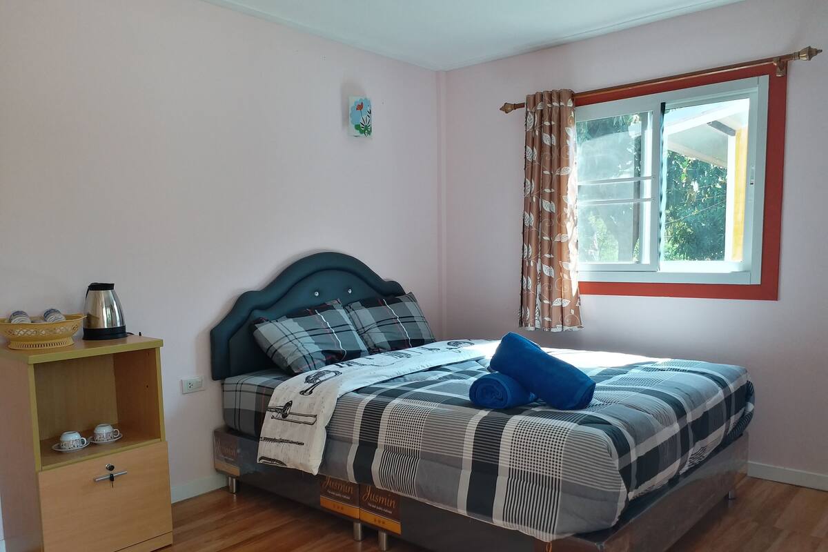 Une chambre bien rangée, avec un lit, une table de chevet sur laquelle se trouve une bouilloire, une petite étagère avec des tasses et une fenêtre avec des rideaux.