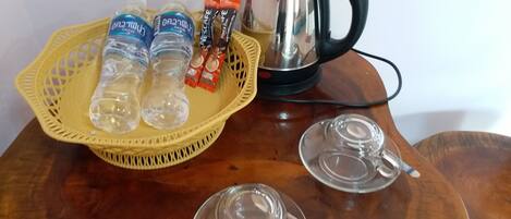 A wooden table with a yellow woven basket containing water bottles and coffee packets, a kettle, and two glass cups with lids.