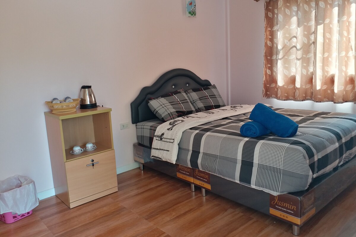 Un dormitorio ordenado con cama, mesita de noche, una pequeña estantería y una ventana con cortinas.