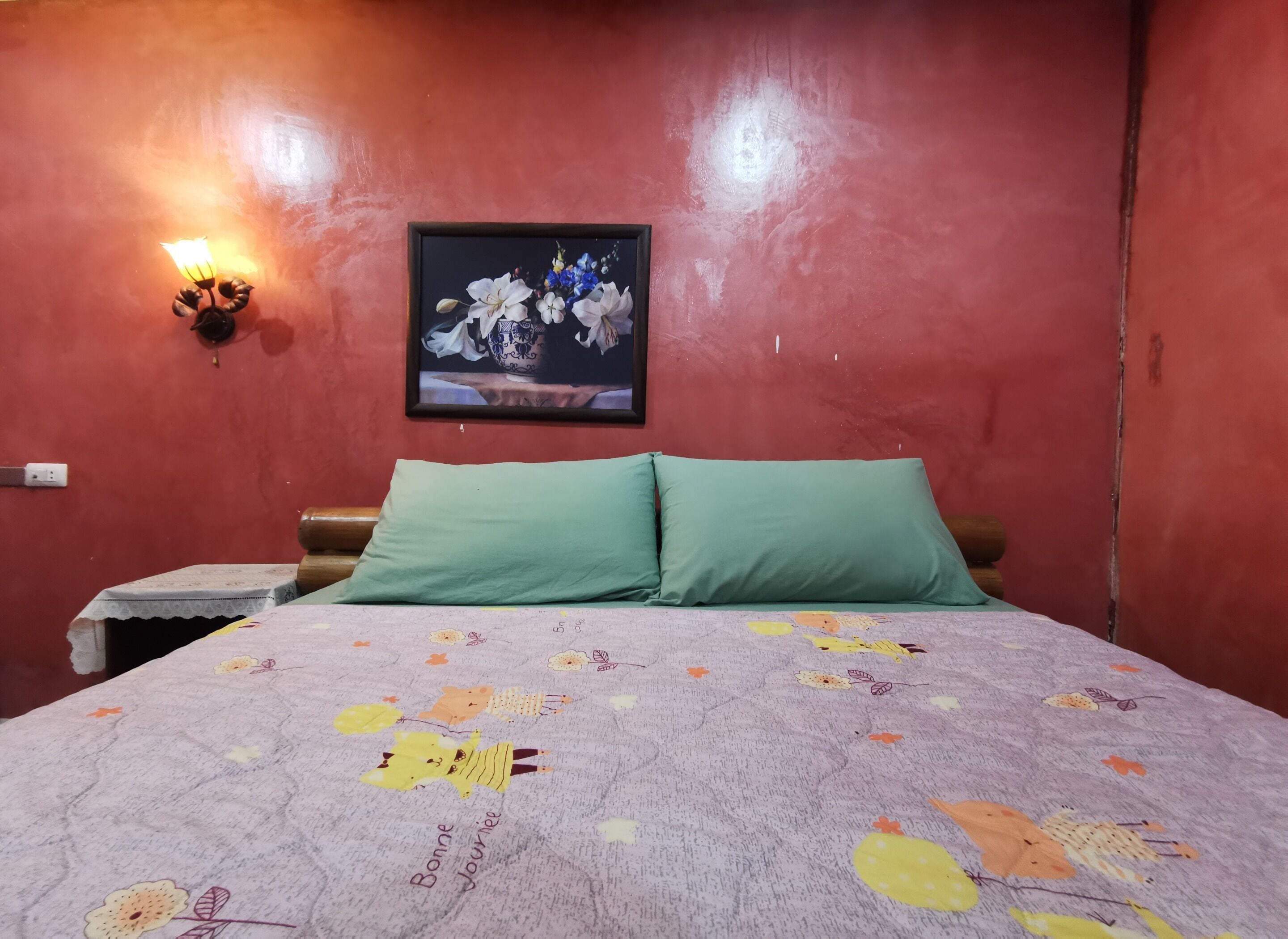 Una habitación con cama, almohadas verdes, una lámpara de pared y una obra de arte floral enmarcada.