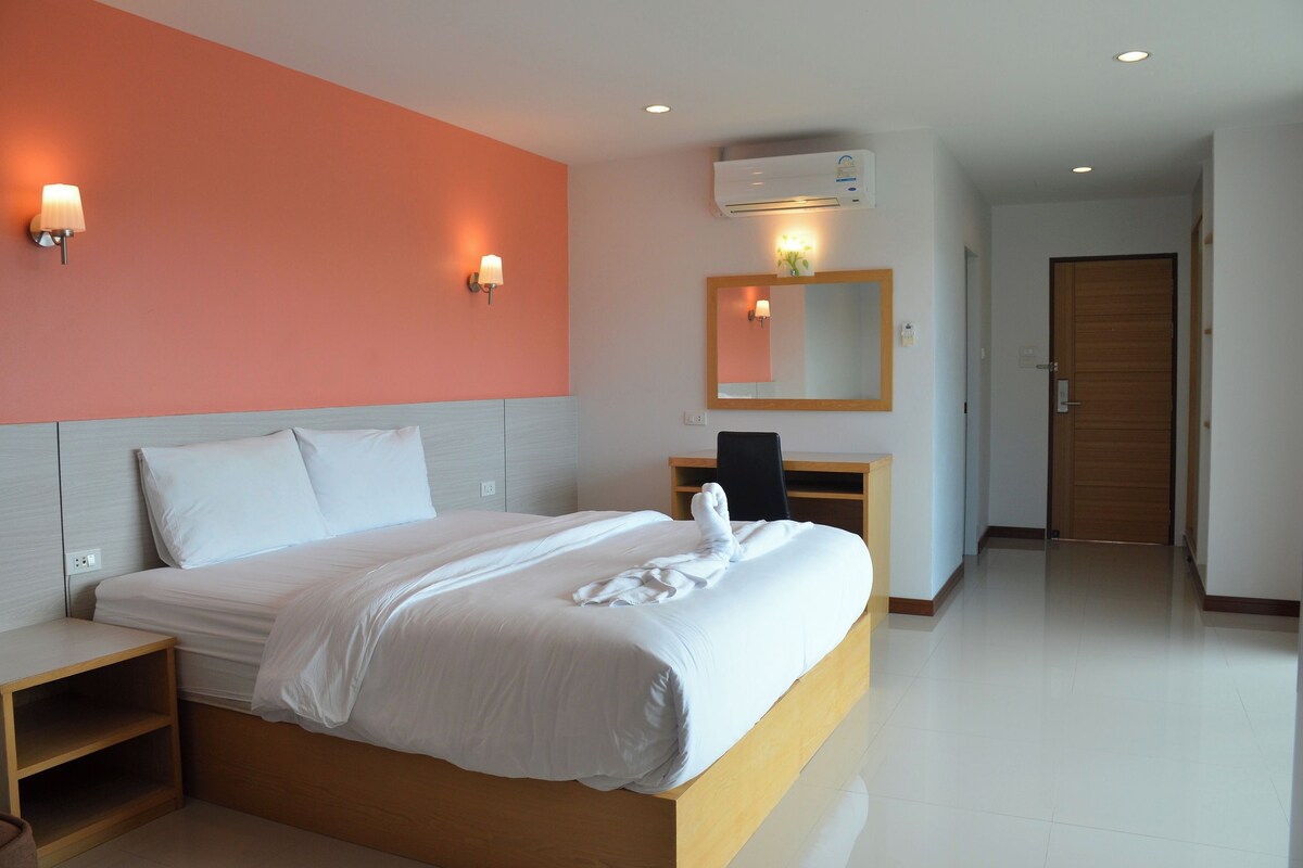 Ein Hotelzimmer mit einem großen Bett, einem Nachttisch aus Holz, einem Schreibtisch mit Stuhl, einem Spiegel und einer Klimaanlage.