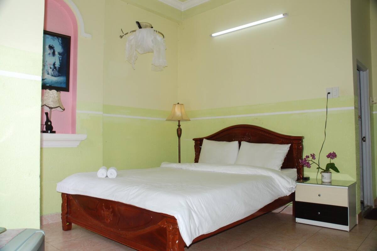 A bedroom with a wooden bed, a nightstand, a lamp, and a wall-mounted shelf with a picture and a white garment.