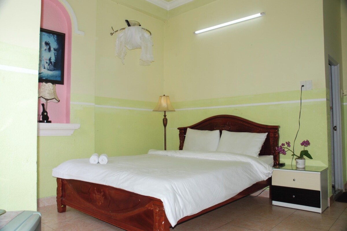 Un dormitorio con cama de madera, una mesita de noche, una lámpara y un estante en la pared con un cuadro y una prenda blanca.