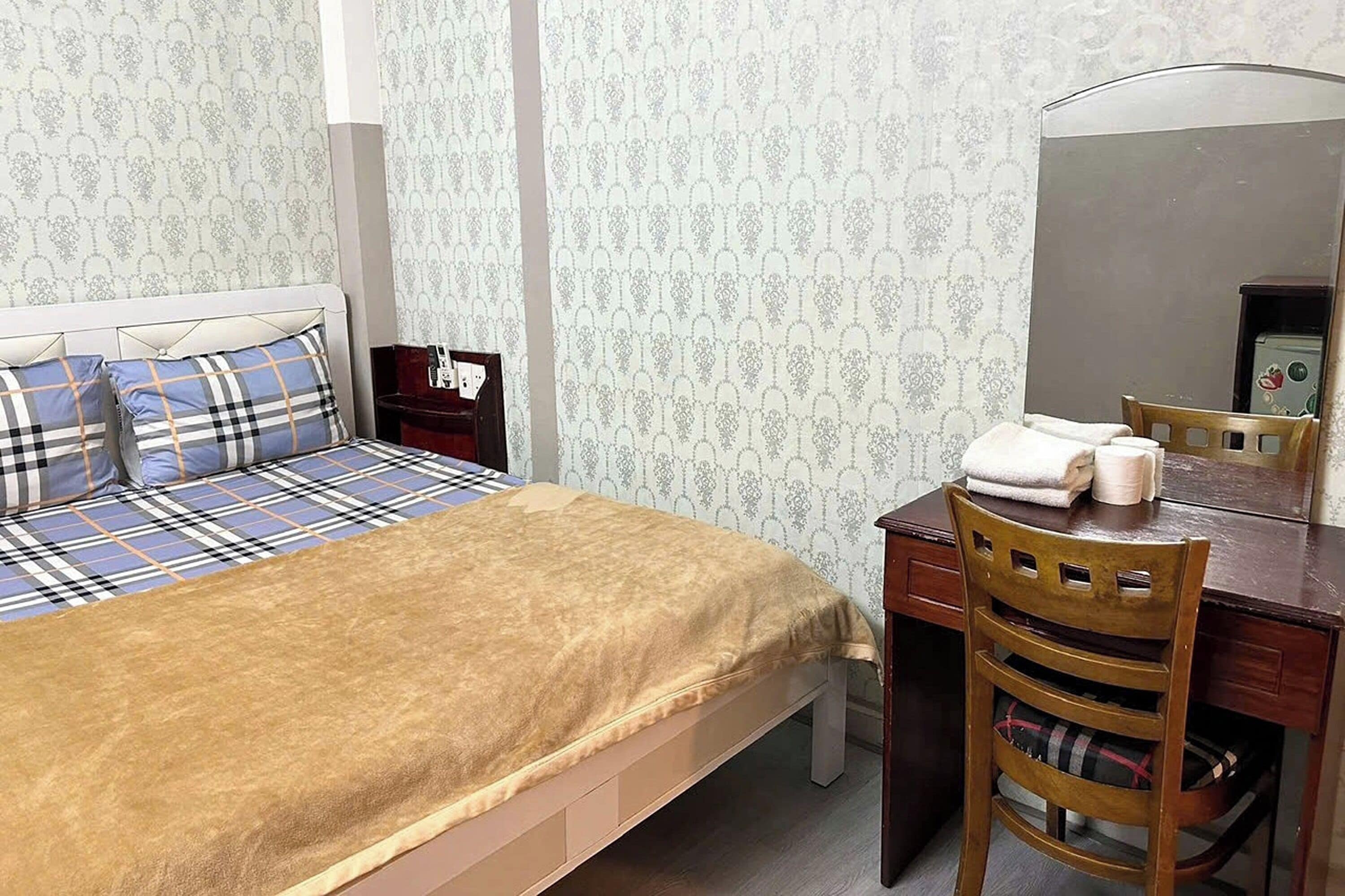 Une chambre à coucher bien rangée, avec un lit, un bureau en bois et une chaise, un miroir et une petite table de chevet avec une lampe.