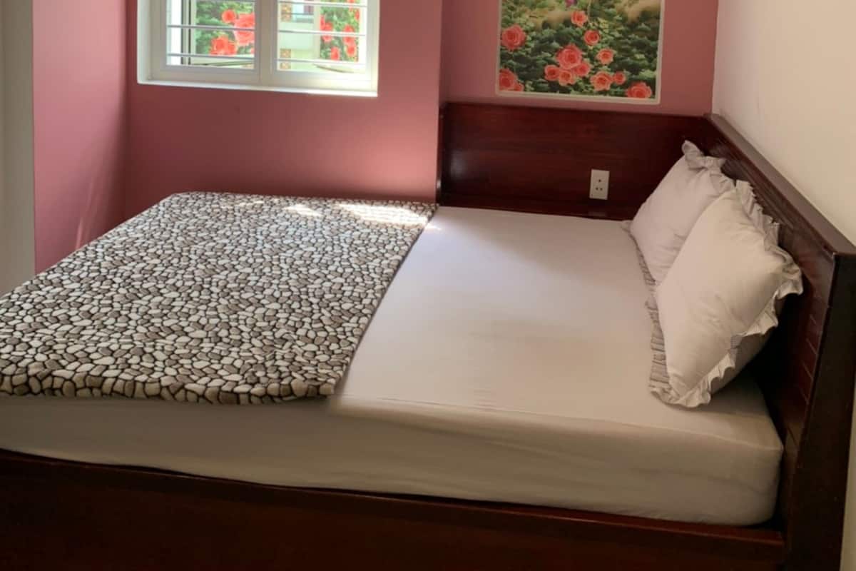 A small bedroom with a bed, a window with a view of a garden, and an air conditioning unit on the wall.