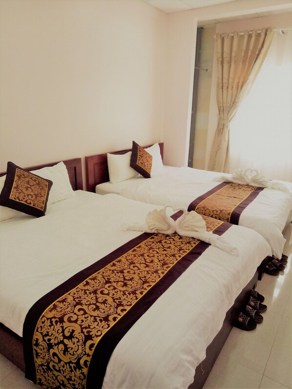 A hotel room with two beds, each with a white bedspread and a decorative gold and brown border. There are pillows with a similar pattern, and a pair of slippers is visible on the floor.