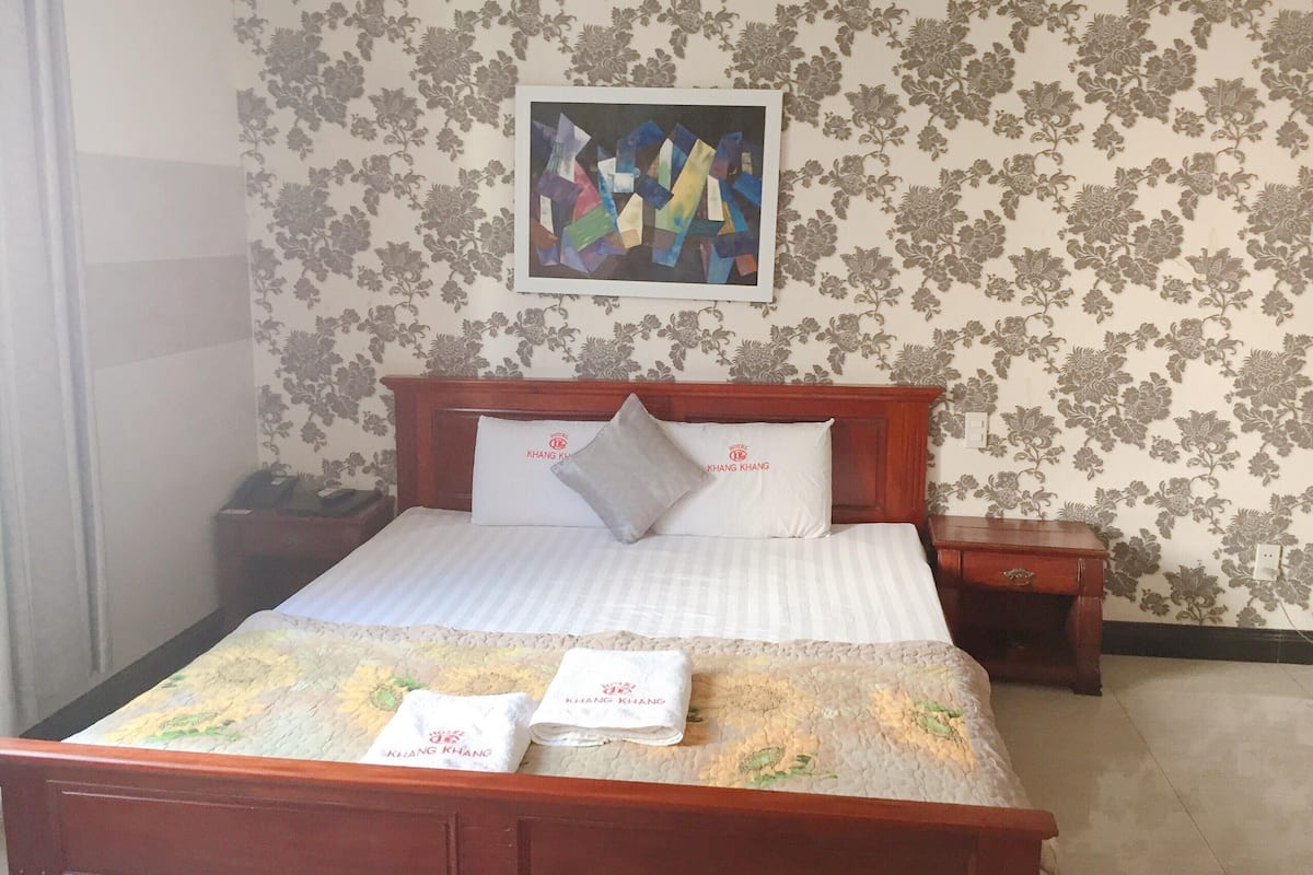 Une chambre d’hôtel avec un lit, deux oreillers, une table de chevet et une commode, le tout agrémenté d’un papier peint à motifs floraux.