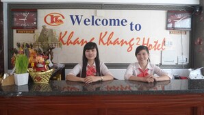 A reception desk at Khans Hotel with a welcome sign, two staff members, and a decorative plant.