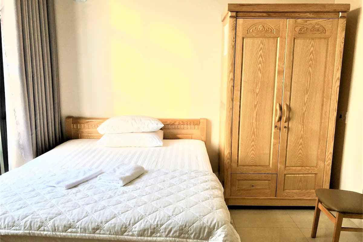 A neatly made bed with a quilted white duvet and two pillows, a wooden wardrobe with two doors, and a wooden chair.
