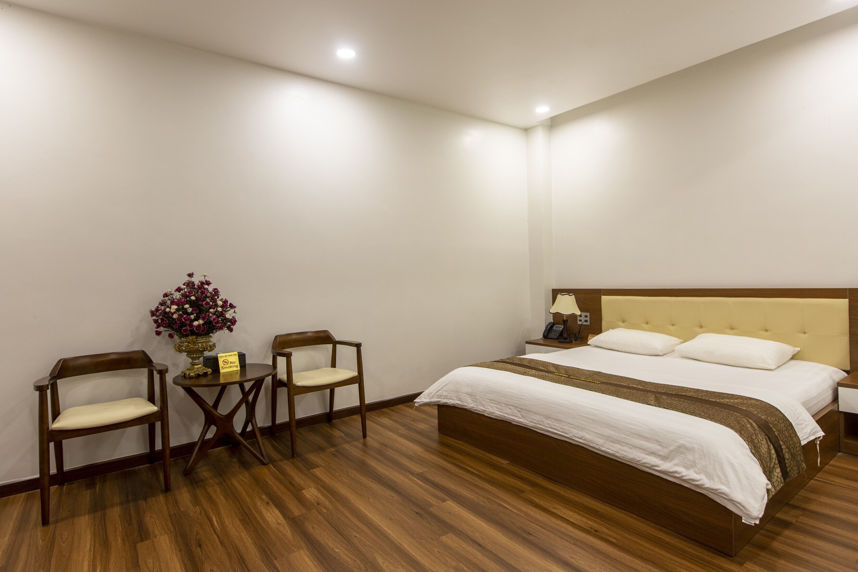 A hotel room with a large bed, two wooden chairs, a small table with a vase of flowers, and a bedside table with a lamp.