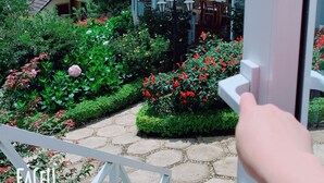 A view of a house with a tiled roof, a garden with red flowers, and a dining area with tables and chairs. - Bungalow Lam Phuong Cac Hill (Xuan Truong - Da Lat)