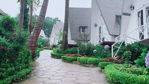 A pathway lined with lush greenery and small flower beds, leading to white buildings with sloped roofs. - Bungalow Lam Phuong Cac Hill (Xuan Truong - Da Lat)