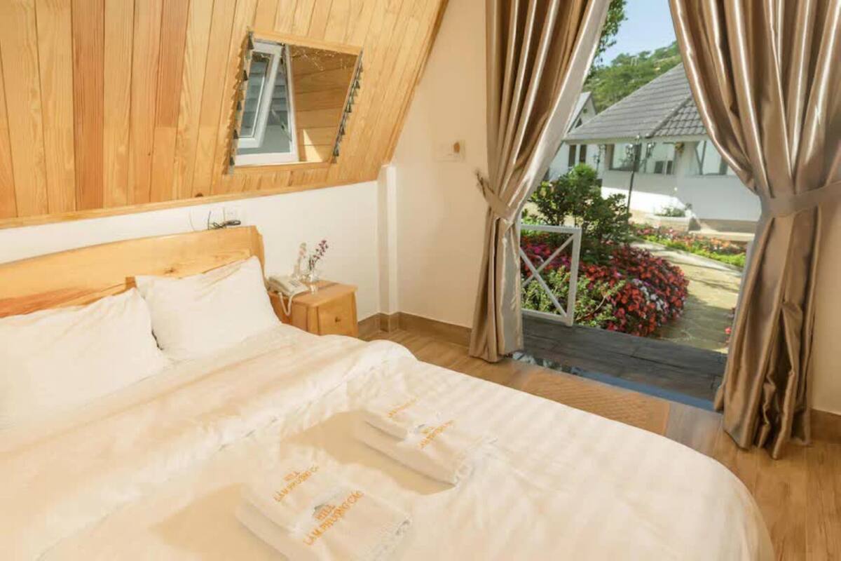 A bedroom with a wooden ceiling, a bed with white linens, a bedside table with a lamp, and a view of a garden through a window.