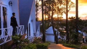 A house with a white porch, a small garden, and a view of trees and a sunset. - Bungalow Lam Phuong Cac Hill (Xuan Truong - Da Lat)