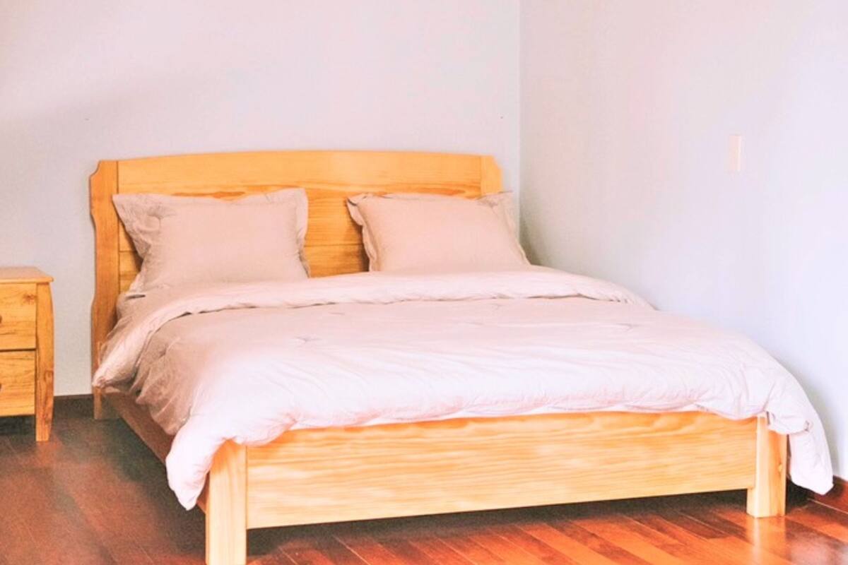 A wooden bed with a headboard, two pillows, and a light-colored blanket in a room with wooden flooring and a white wall.