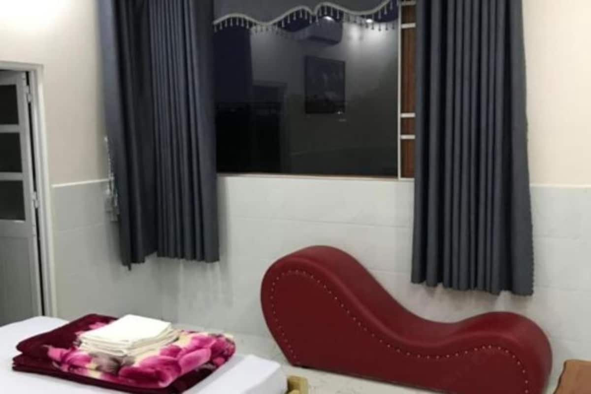 Ein Hotelzimmer mit Bett, rotem, gepolsterten Kopfteil, Fenster mit Vorhängen und einem kleinen Tisch mit einem Stapel weißer Handtücher.