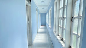 Un long couloir étroit, avec de grandes fenêtres d’un côté et des portes de l’autre, menant à une pièce au bout.