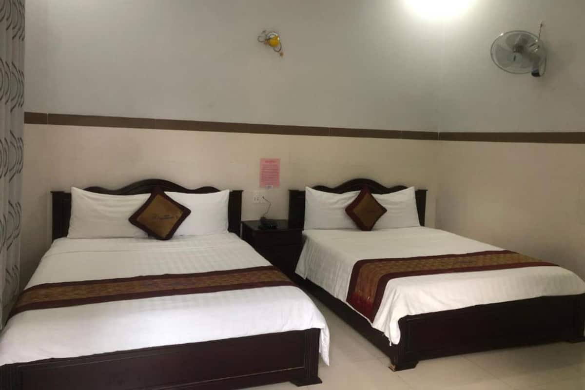 Ein Hotelzimmer mit zwei Einzelbetten, jeweils mit einem hölzernen Kopfteil, weißer Bettwäsche und einem braunen Zierkissen. Es gibt einen Nachttisch mit einer Lampe und einen Wandventilator.