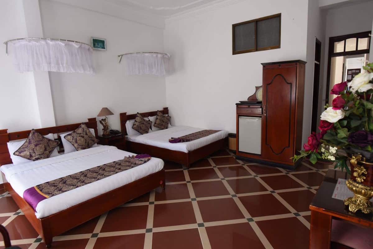 A hotel room with two wooden beds, a wooden wardrobe, a small table with a vase of flowers, and a tiled floor.