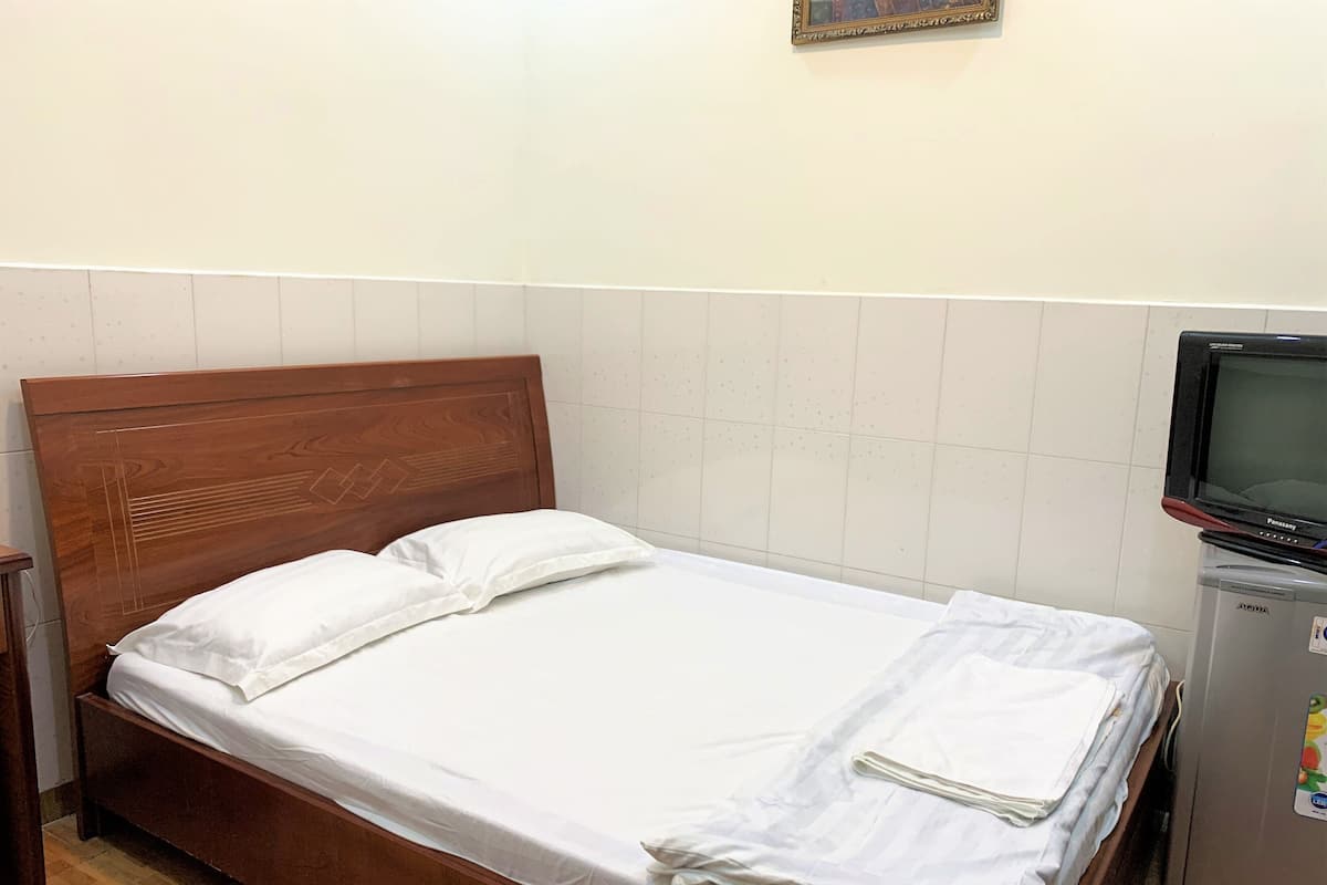 Un lit simple avec du linge de lit blanc et des oreillers, une tête de lit en bois, une petite télévision posée sur un meuble et un réfrigérateur dans un coin.