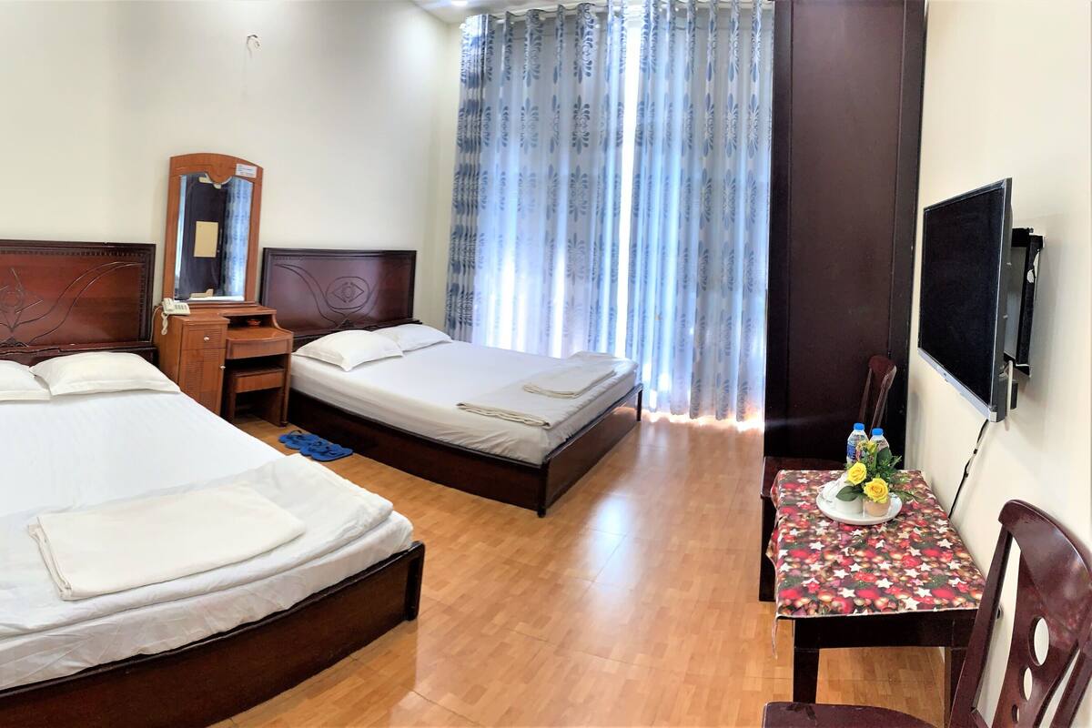 Une chambre d’hôtel avec deux lits, un bureau en bois, un miroir, une télévision et une petite table recouverte d’une nappe à motifs floraux.